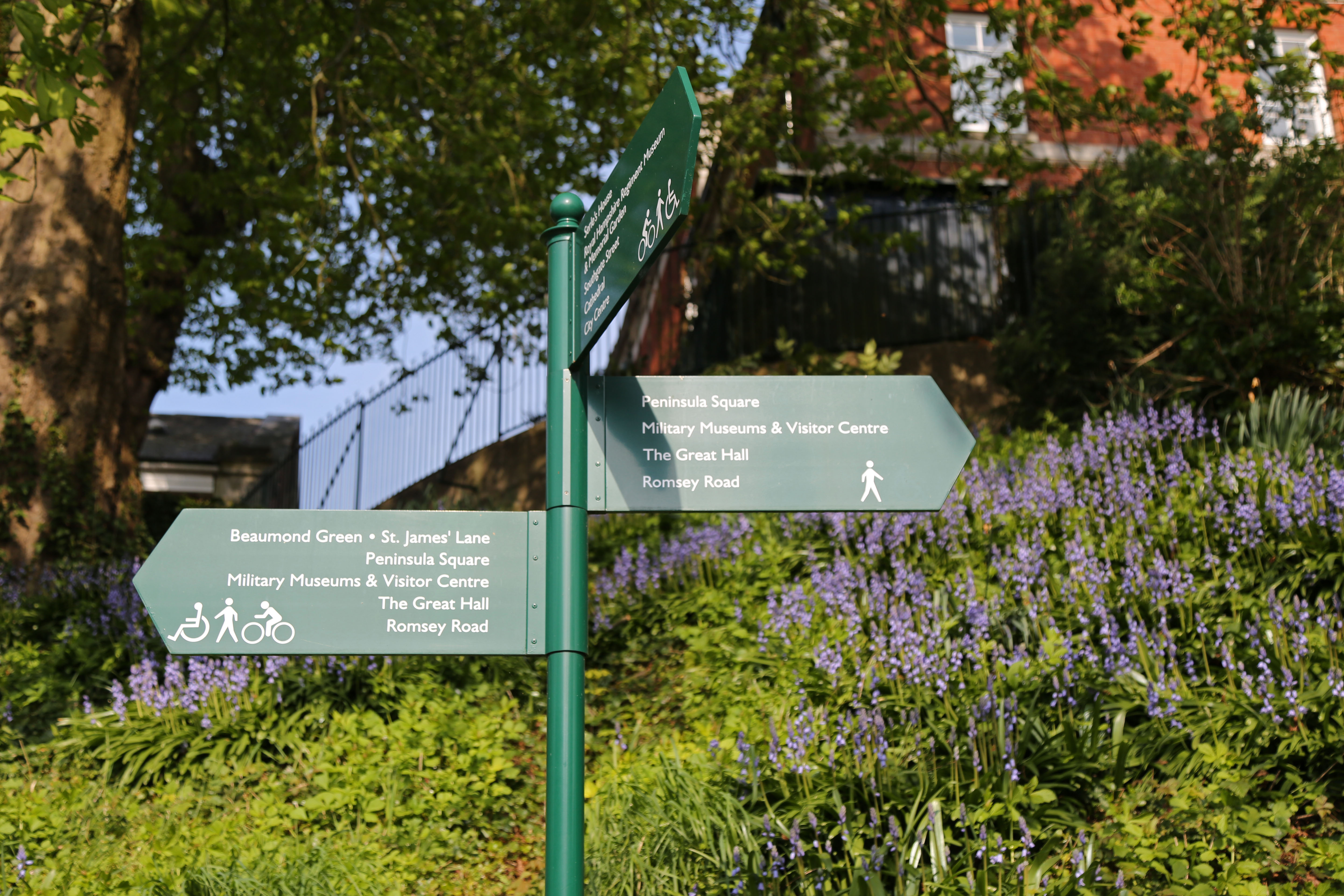 Three signs pointing to the historical landmarks at Winchester Barracks, with blue flowers in the background