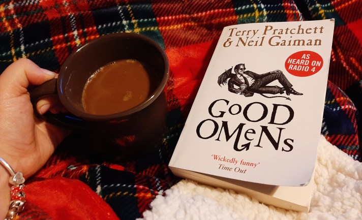 Rosie's hand holding a cup of hot chocolate, next to a copy of the novel 'Good Omens'. The background is a red checkered blanket.