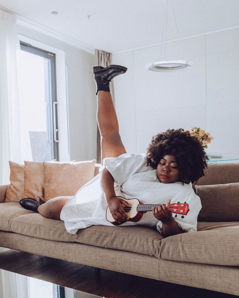 Stephanie laying on a sofa, wearing a white dress. he leg is in the air and she is playing the ukulele