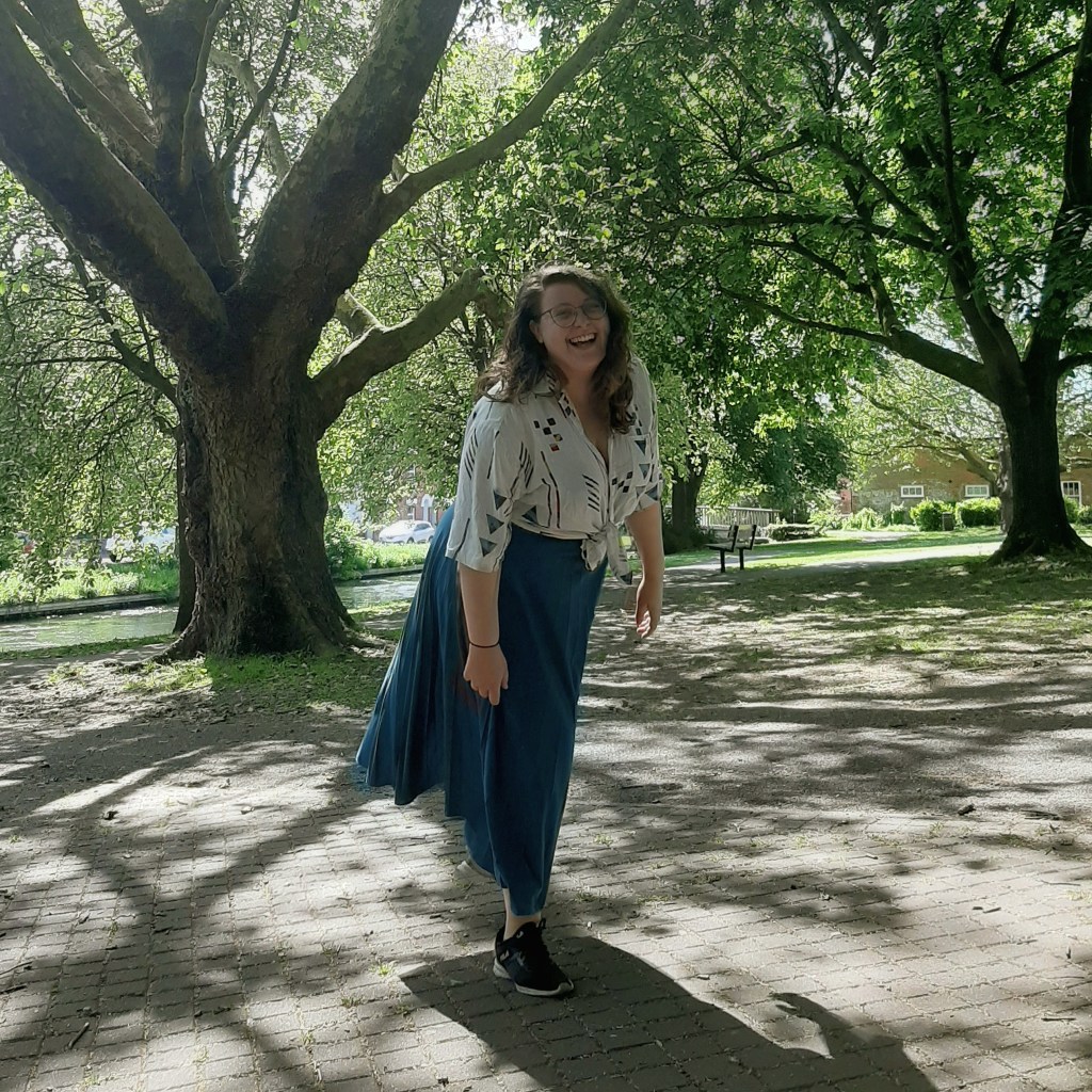 Rosie wearing a blue skirt, and a whiteshirt (with colourful shapes on it). She is smiling at the camera, with a background of green grass and trees