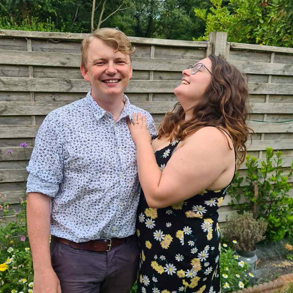 Rory and ROsie stood next to each other. Rosie is laughing, and has her left hand on his chest, showcasing the ring