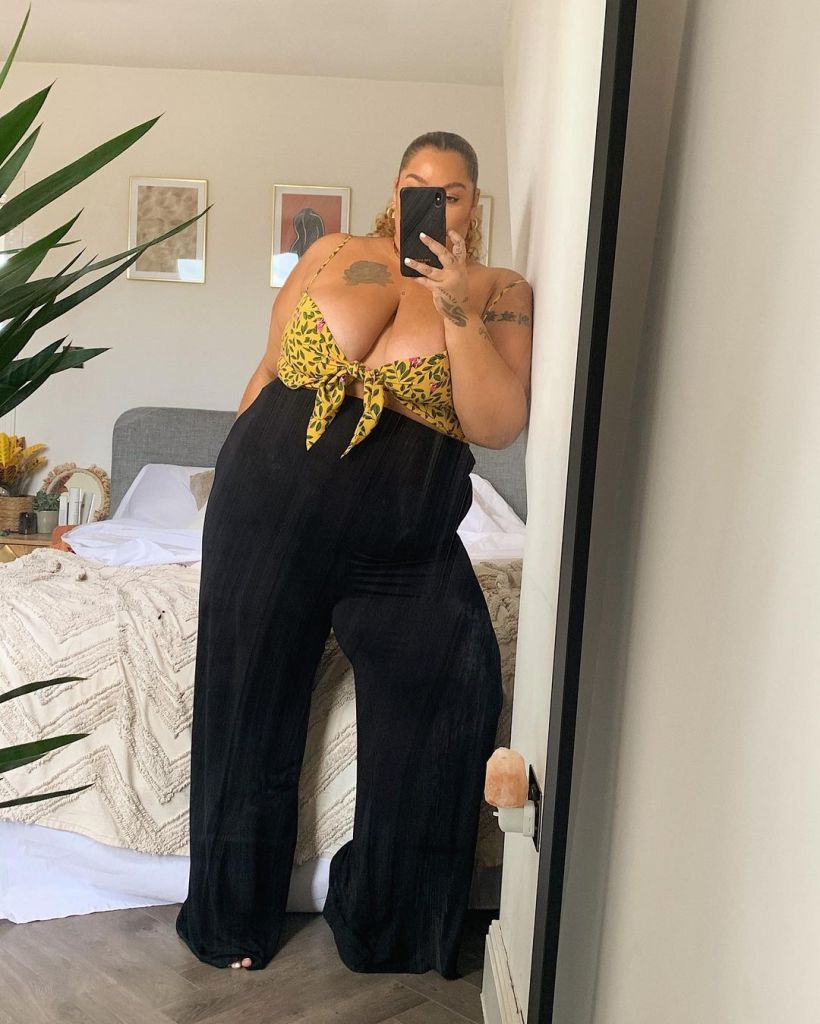 Grace is stood leaning against the wall, taking a mirror selfie. She is wearing black trousers, and a yellow floral printed bralet style top. Her face is obscured by the camera.