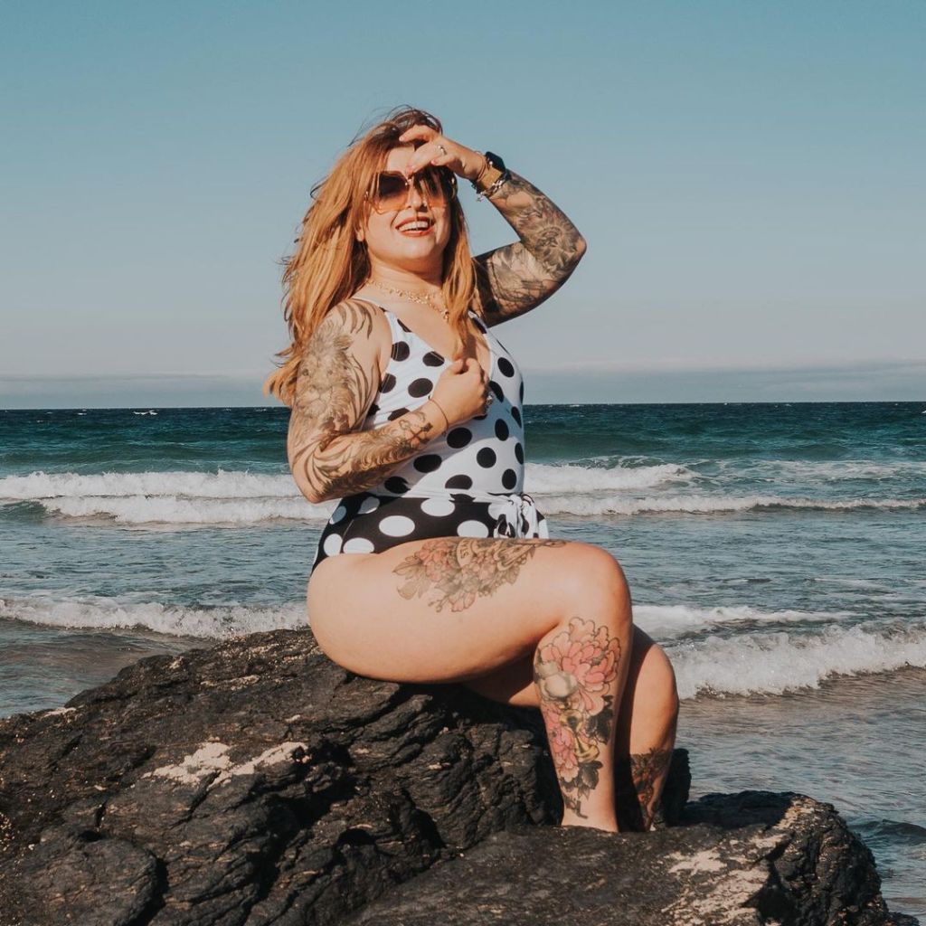 Helen is sat on a rock with the sea behind her, and she is looking at the sunglasses. She is wearing a black and white swimsuit, sunglasses, and her tattoos on her legs are popping.