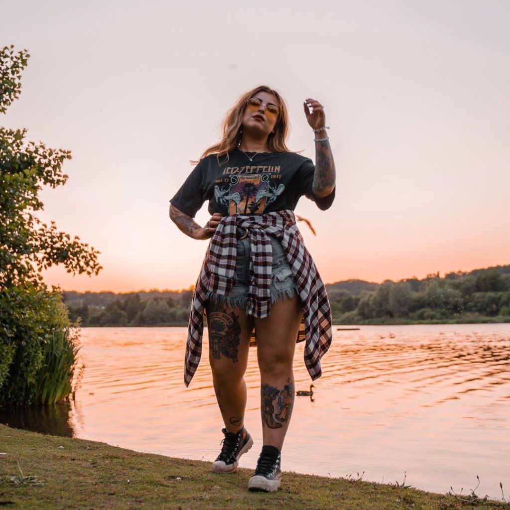 Helen is walking towards the camera, wearing a Led Zeppelin tee, denim shorts, and a check shirt around her waist. The sun is setting on a lake behind her