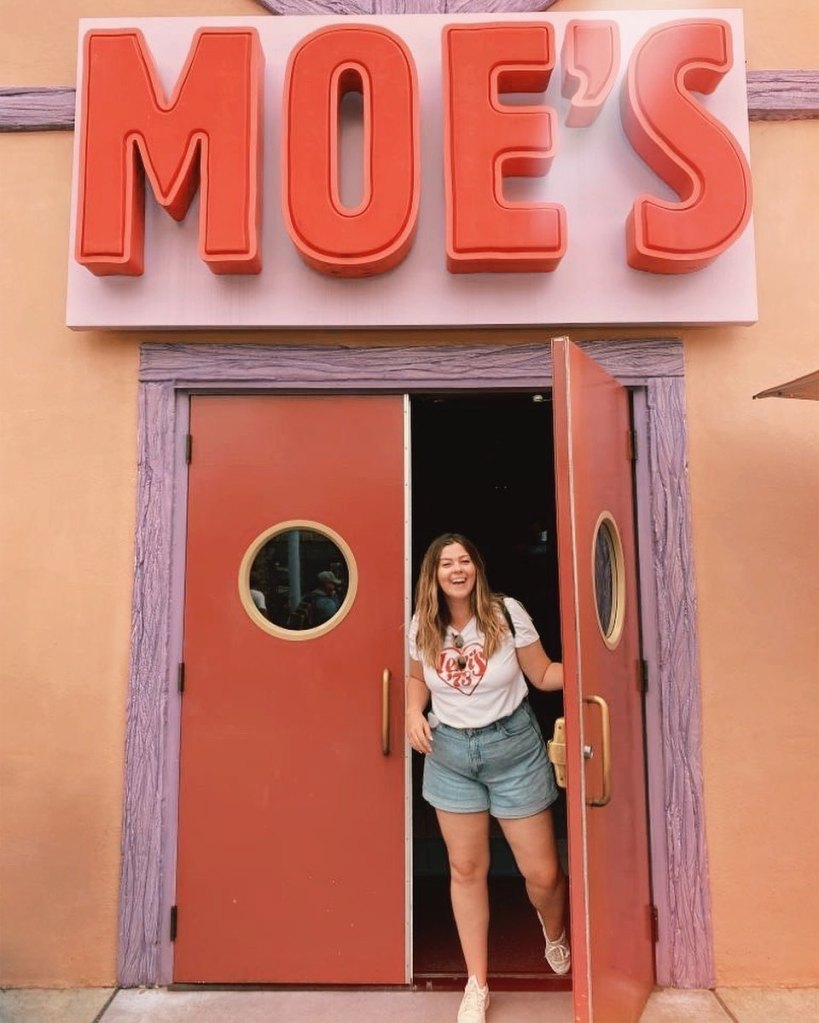 Lucy is posing in the doorway of Moe's Tavern in Universal Studios. She is wearing a white tee, denim shorts, and is smiling at the camera.