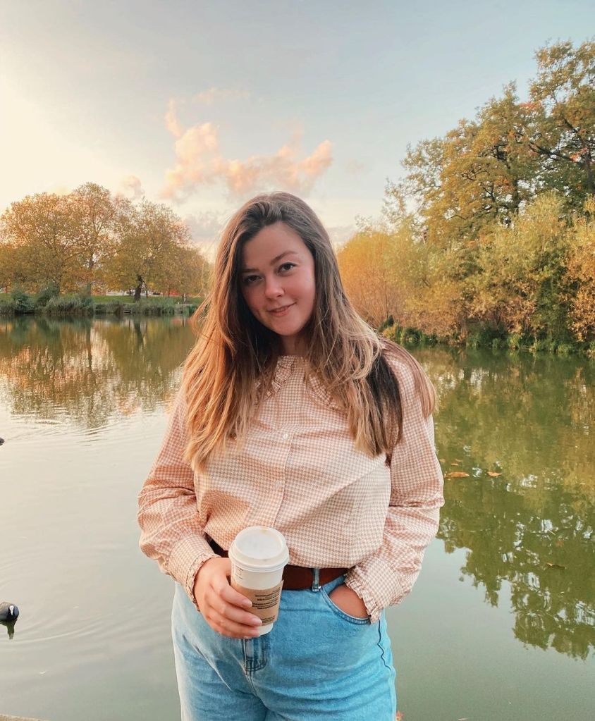 Lucy is wearing a pale pink check shirt, and blue mom jeans. She is posing in front of an autumn lake scene and looking directly at the camera.