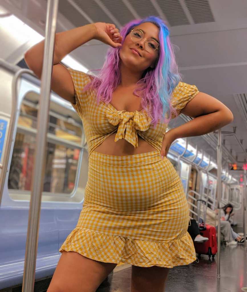 Megan is wearing a yellow check mini dress, with a cut out in the middle. She has pink and purple hair, is looking directly at the camera, and is posing on a train.