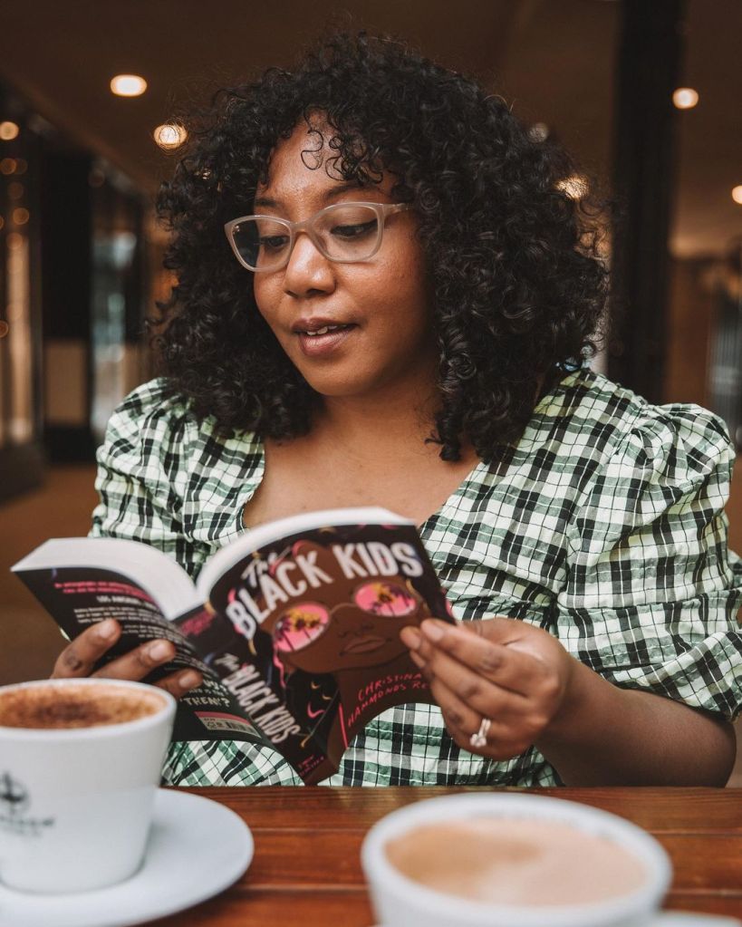 Nicole is wearing a black and white check puff sleeve top, and tan glasses. She is reading a novel called 'The Black Kids', and there's coffee in the foreground