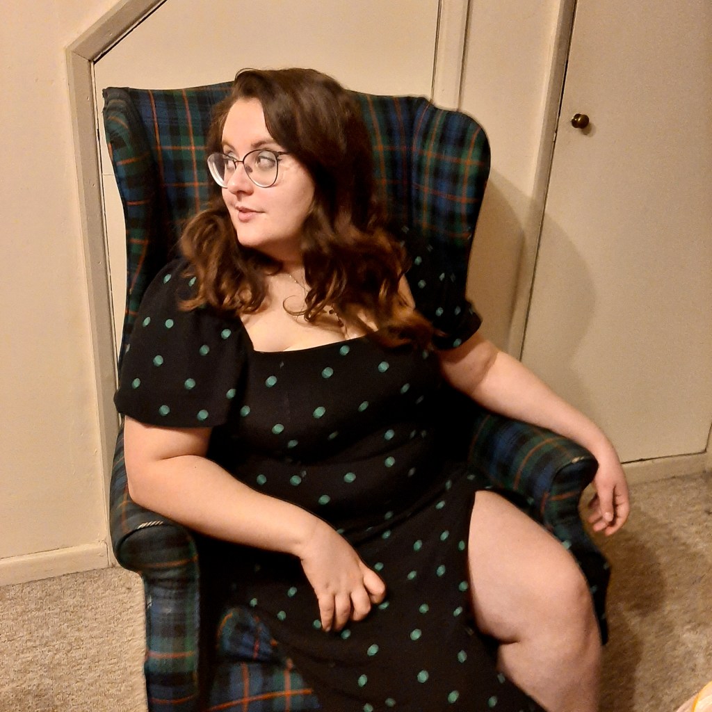 Rosie looking at the camera. Her hair is brown, and she is wearing a black dress with green spots. She is sitting on a green and blue armchair, and is looking to the right. It's an almost full body shot.