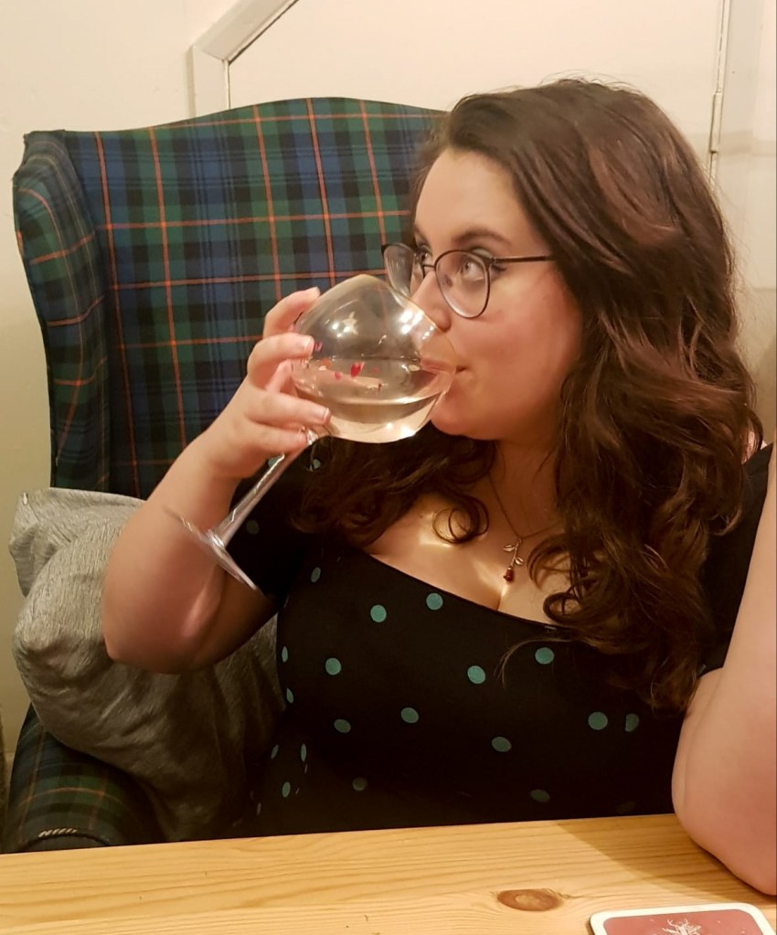 Rosie looking at the camera. Her hair is brown, and she is wearing a black dress with green spots. She is sitting on a green and blue armchair, and is looking to the right. She is holding a large gin glass to her lips.