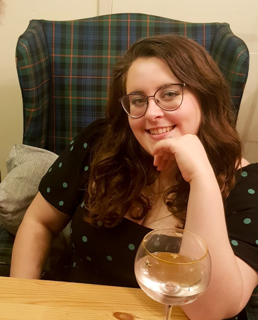 Rosie looking at the camera. Her hair is brown, and she is wearing a black dress with green spots. She is sitting on a green and blue armchair, and is leaning on her left hand. There's a large gin glass in front of her.