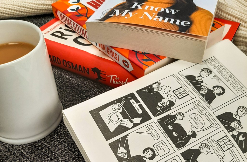White mug filled with milky tea. Three red/orange toned books in the background. Black and white graphic novel lays open.