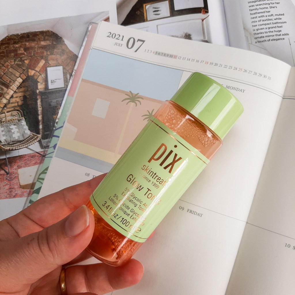 A pink and green bottle of Pixi's Glow Tonic, against a background of a cream diary and white magazine