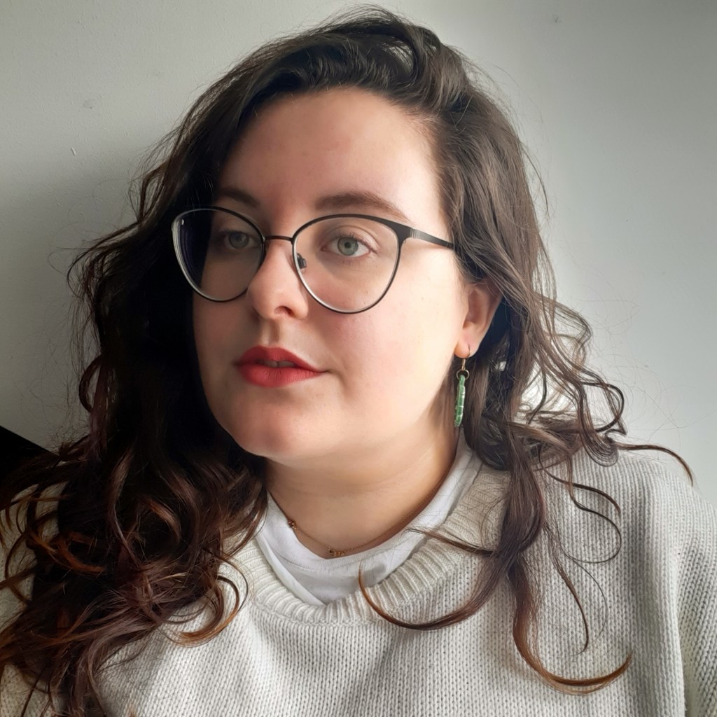 A picture of Rosie looking to the left. She is wearing a light grey jumper, has brown hair, and black glasses.