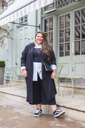 Callie is wearing a white shirt, black jumper, black coat black culottes, and black and white trainers. Callie has brown hair and is a size 22 woman.