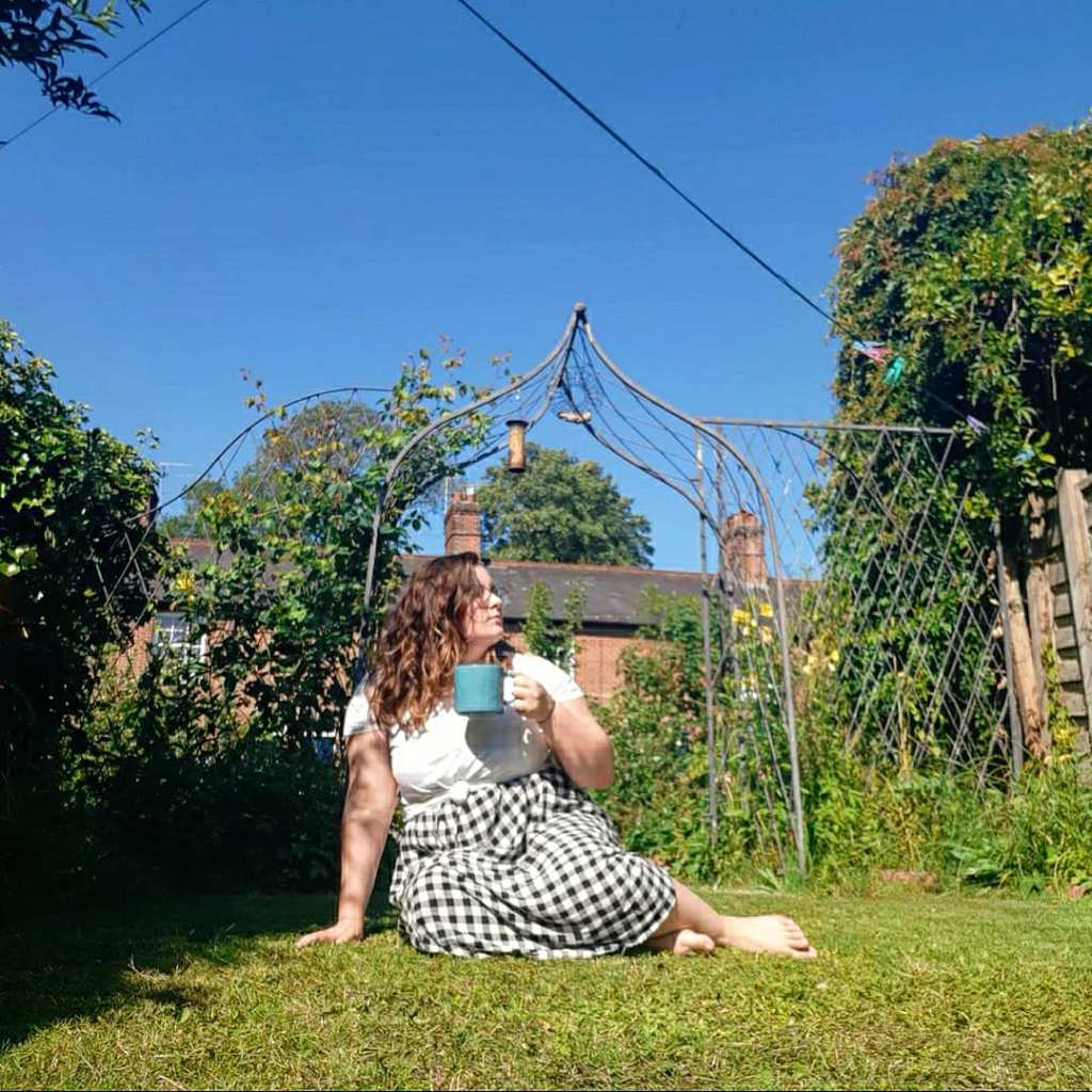 Rosie is sat on green grass, holding a blue mug, and is looking to the left. She is wearing a white tee and black and white checked midi skirt