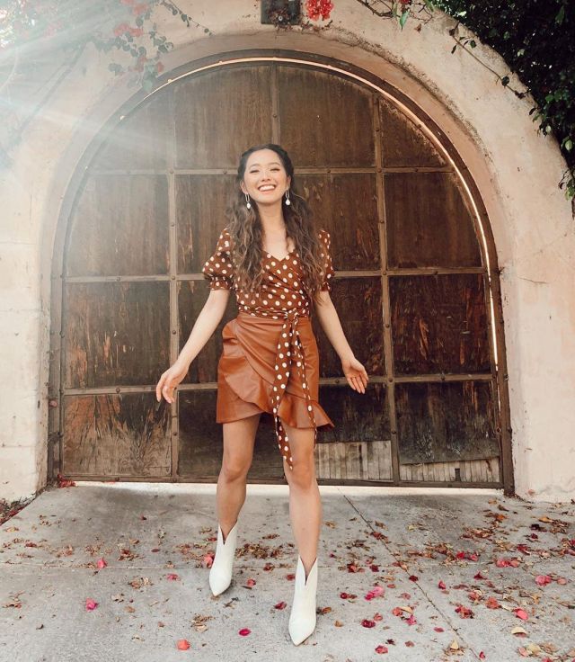 A picture of Jenn Im in front of a brown door. She is wearing a dark orange spotty top, with an orange mini skirt, and white boots. Jenn is a slim Korean American woman with brown hair