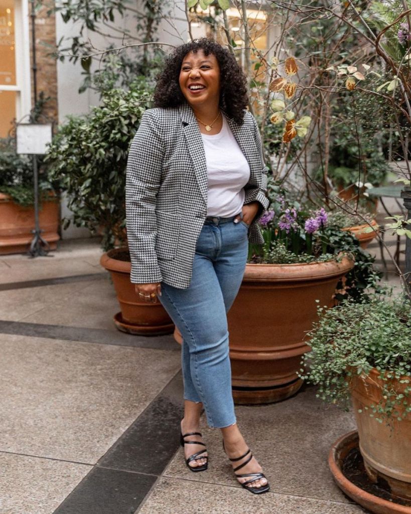 Nicole is wearing a grey check blazer, white tee, blue jeans, and black strappy heels. Nicole has dark hair, and is a mixed-raced, American, size 16 woman.