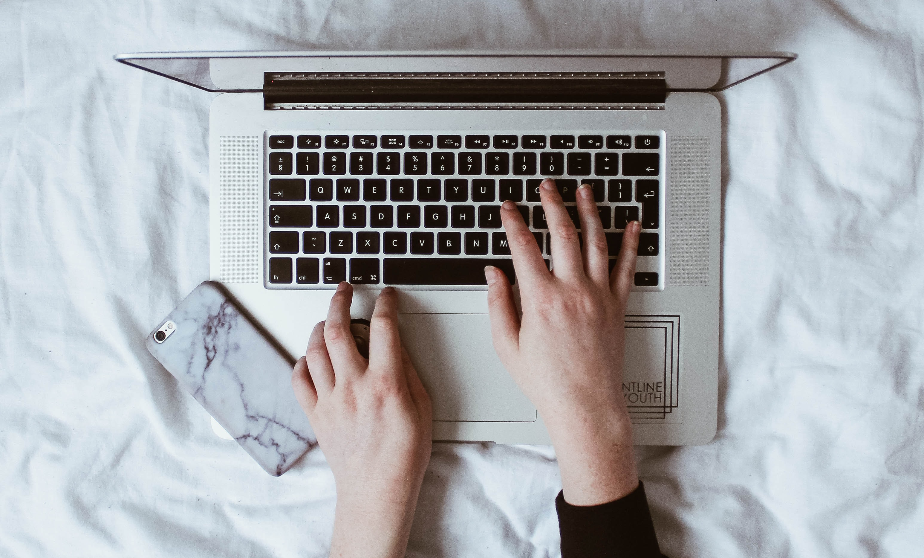 Silver laptop on a white bedsheet background. Two hands are typing. There is a mobile phone in a marble style phone case to the