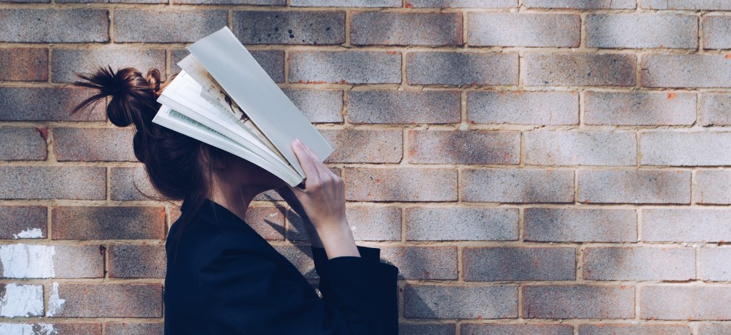 A girl with a messy bun and black blazer, hitting herself in the face with a stack of paper. The brick wall behind her is grey.
