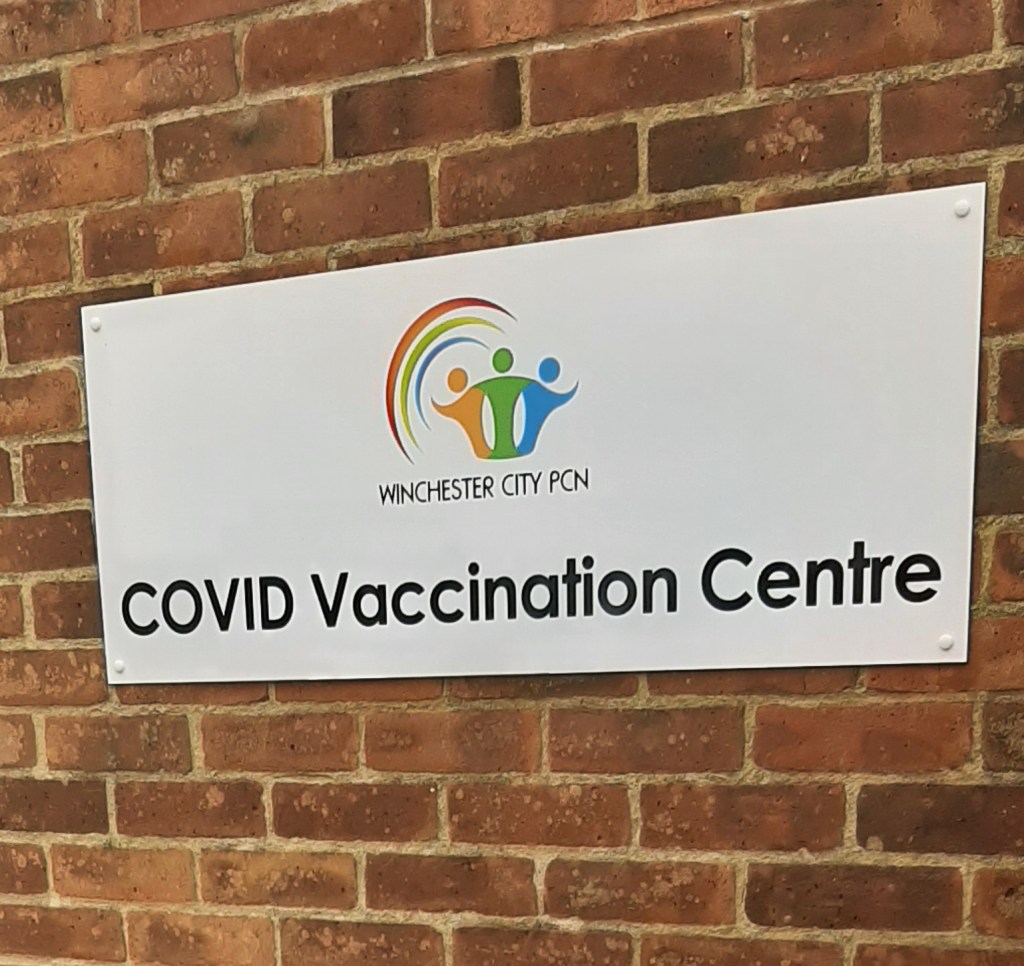 A red brick wall, with a white sign that says 'COVID Vaccination Centre' on it in black text.