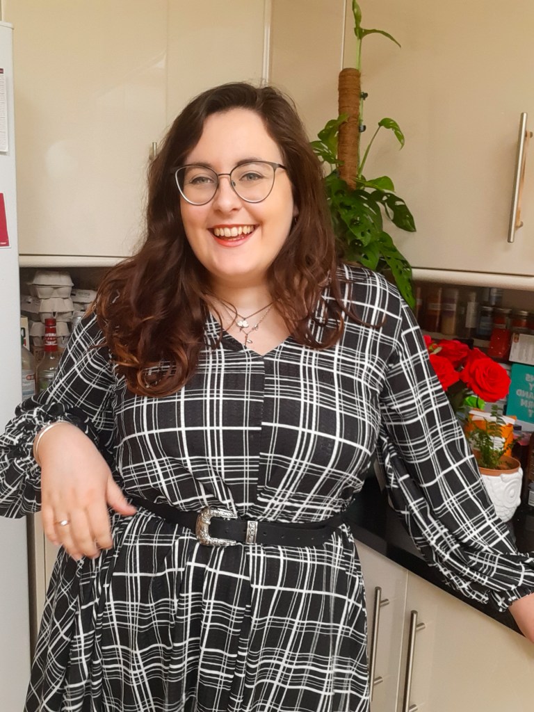 Rosie is laughing, looking at the camera, with her right hand above her hip. She is wearing a long sleeve black dress with white grid marks on it. She is wearing a black belt and has glasses.