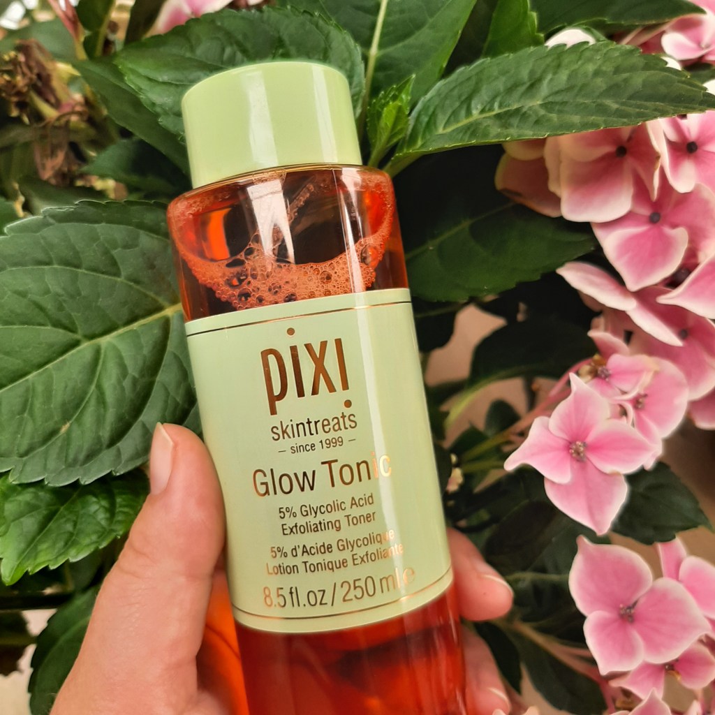Green and pink toner bottle that says 'pikxi skintreats Glow Tonic'. It is held up by Rosie's caucasian hand against green leaves and pink flowers
