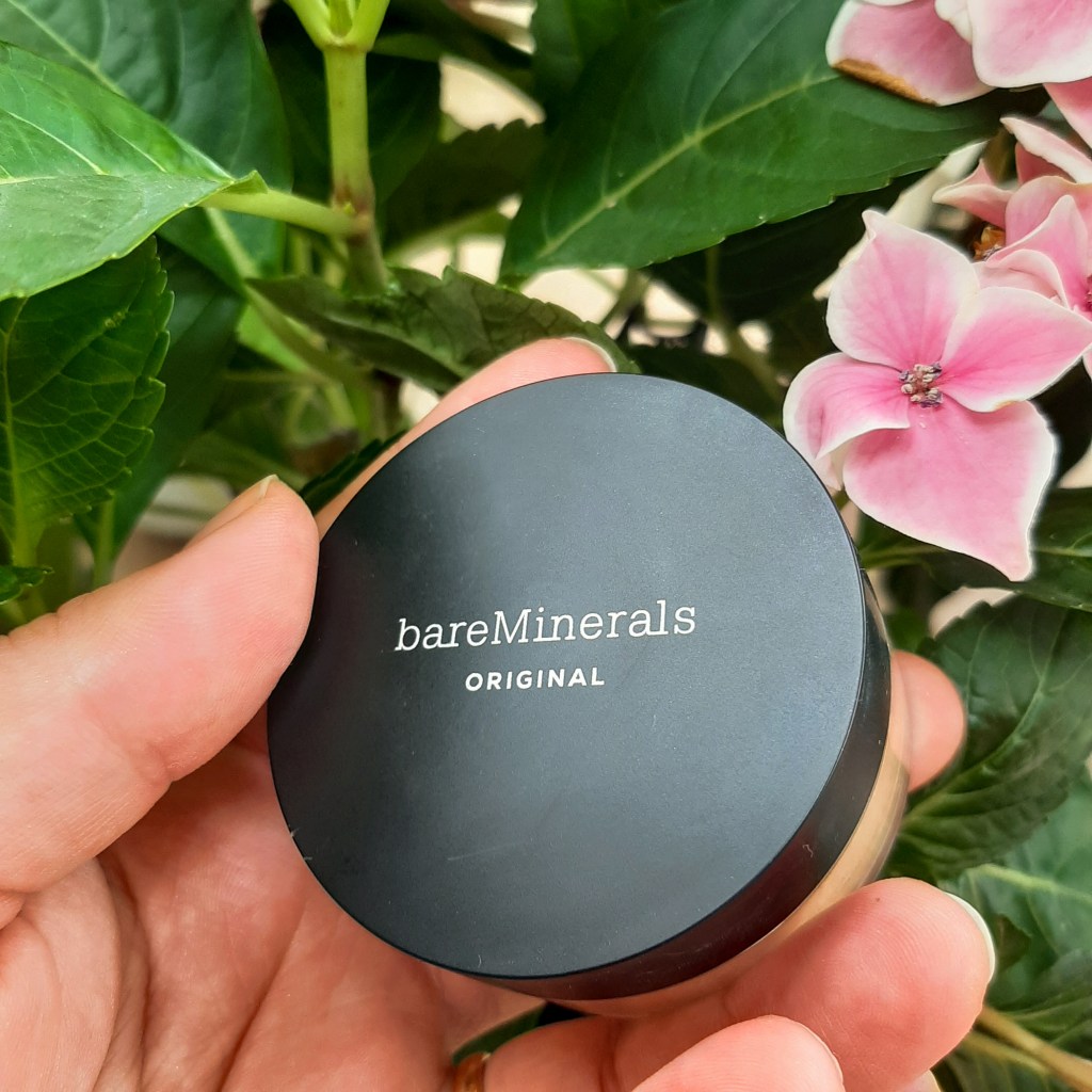 Round foundation container with a black lid that says 'bareMinerals original'. It is held up by Rosie's Caucasian hand against green leaves and pink flowers