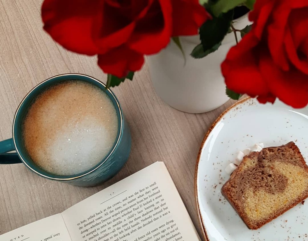 A frothy coffee in a teal mug, next to a white and brown slice of cake. There are red roses