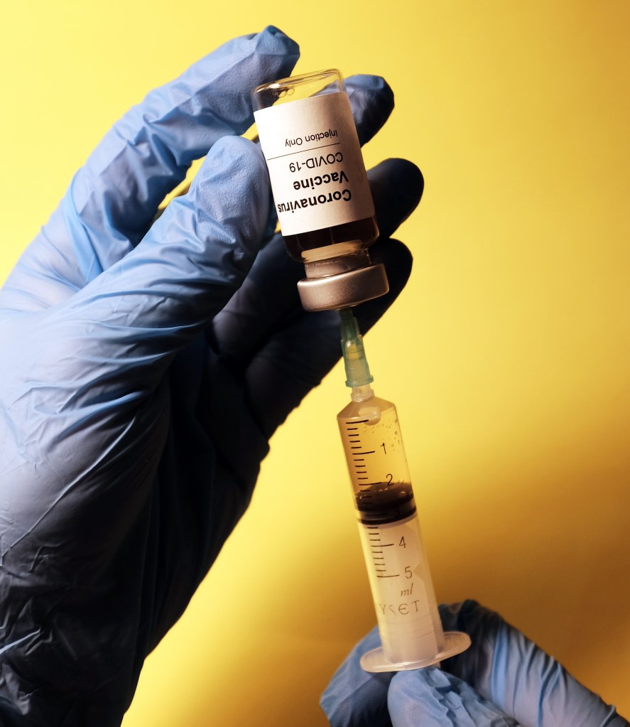 Blue gloved hands holding an injection, which has a glass container on top. The background is yellow.