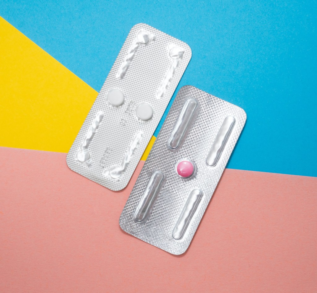 Two silver packets - one with a singular pink pill inside, and the one on th eleft has two white pills inside. They are on a background of pink, yellow, and blue card.