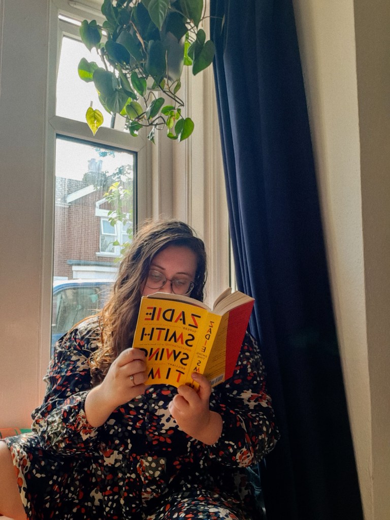 Rosie is sitting in a white window nook, next to a navy curtain. She has dark hair, glasses, and is looking down at a book. She is holding the yellow book in her hands - it is Zadie Smith's Swing Time.