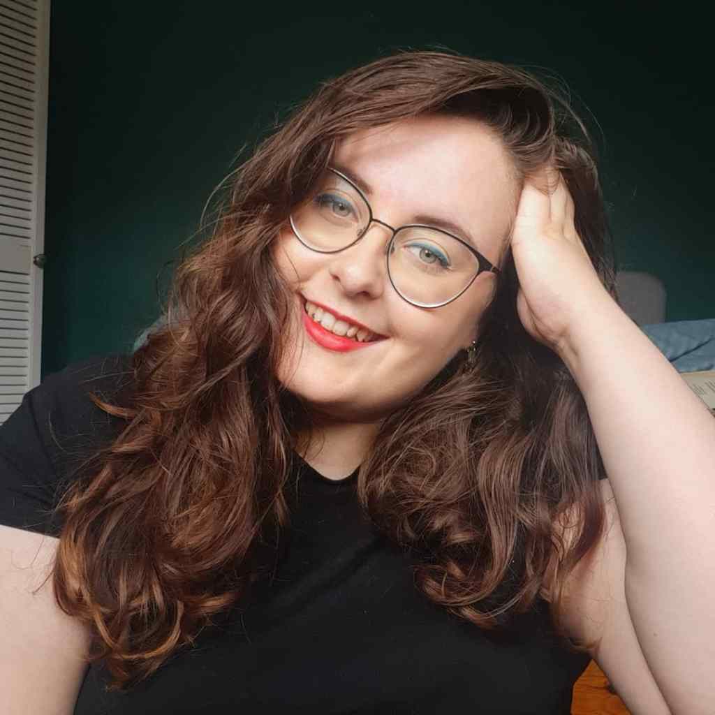 Rosie, a caucasian woman with brunette wavy hair, is looking at the camera with a hand in her hair. She is wearing a black t-shirt, had black glasses, red lips, and blue eyeliner