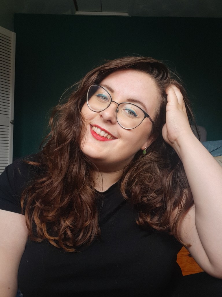 Rosie looking straight at the camera. She has a hand in her hair, wear lips, glasses, and a blue cat eye liner