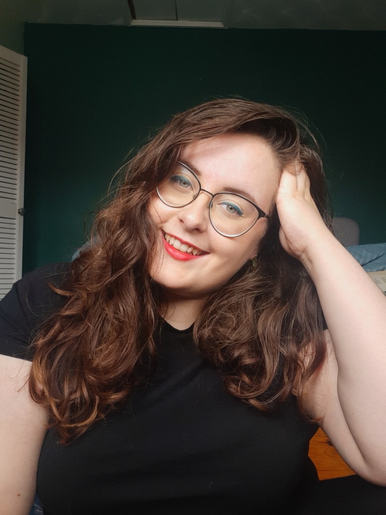 Rosie looking straight at the camera. She has a hand in her hair, wear lips, glasses, and a blue cat eye liner