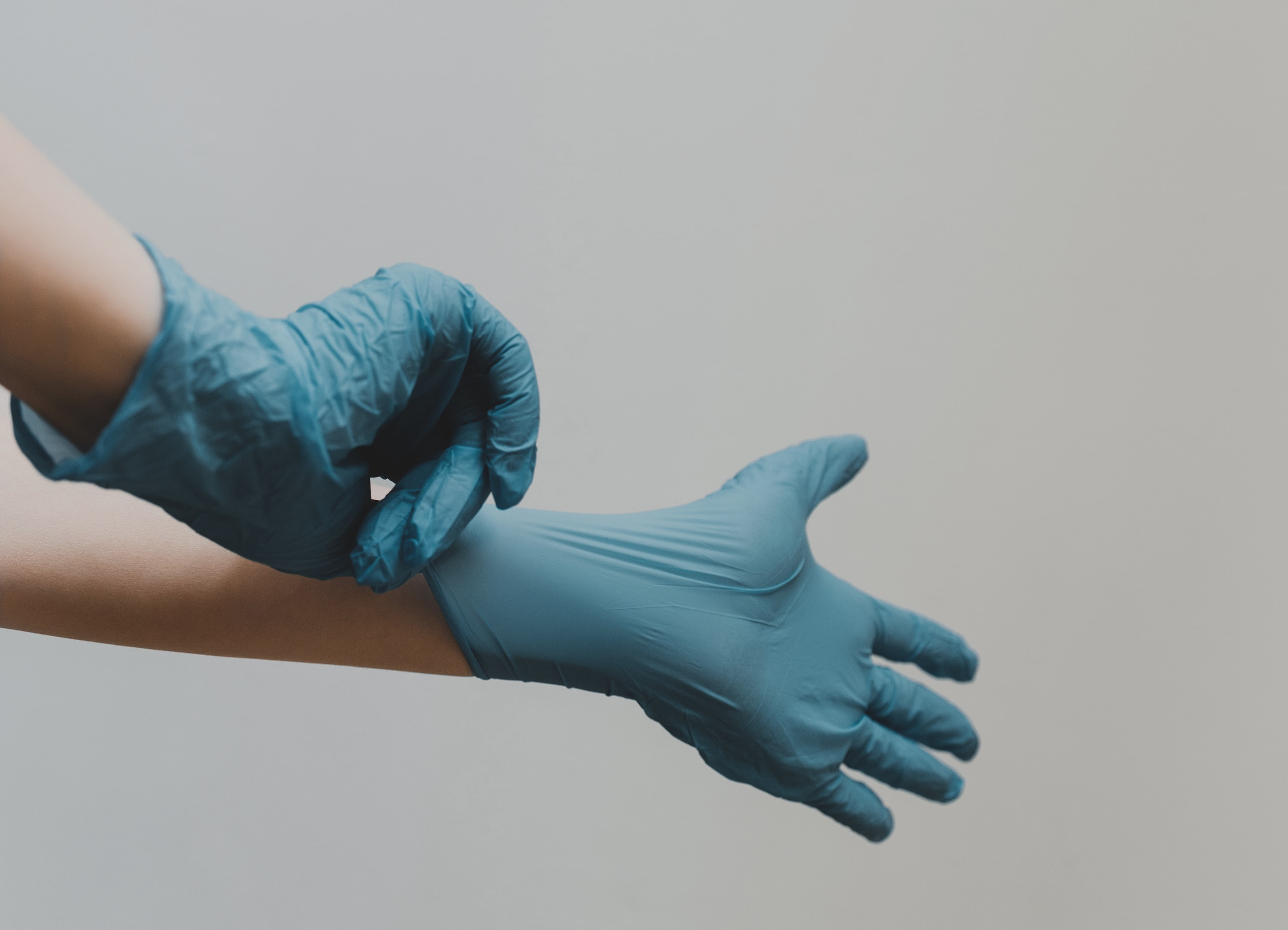 A pair of Caucasian hands pulling on blue medical gloves. The background is off white