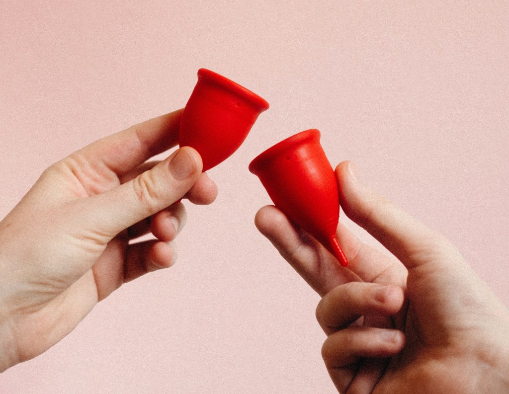 Two hands holding two red period cups against a pale pink background