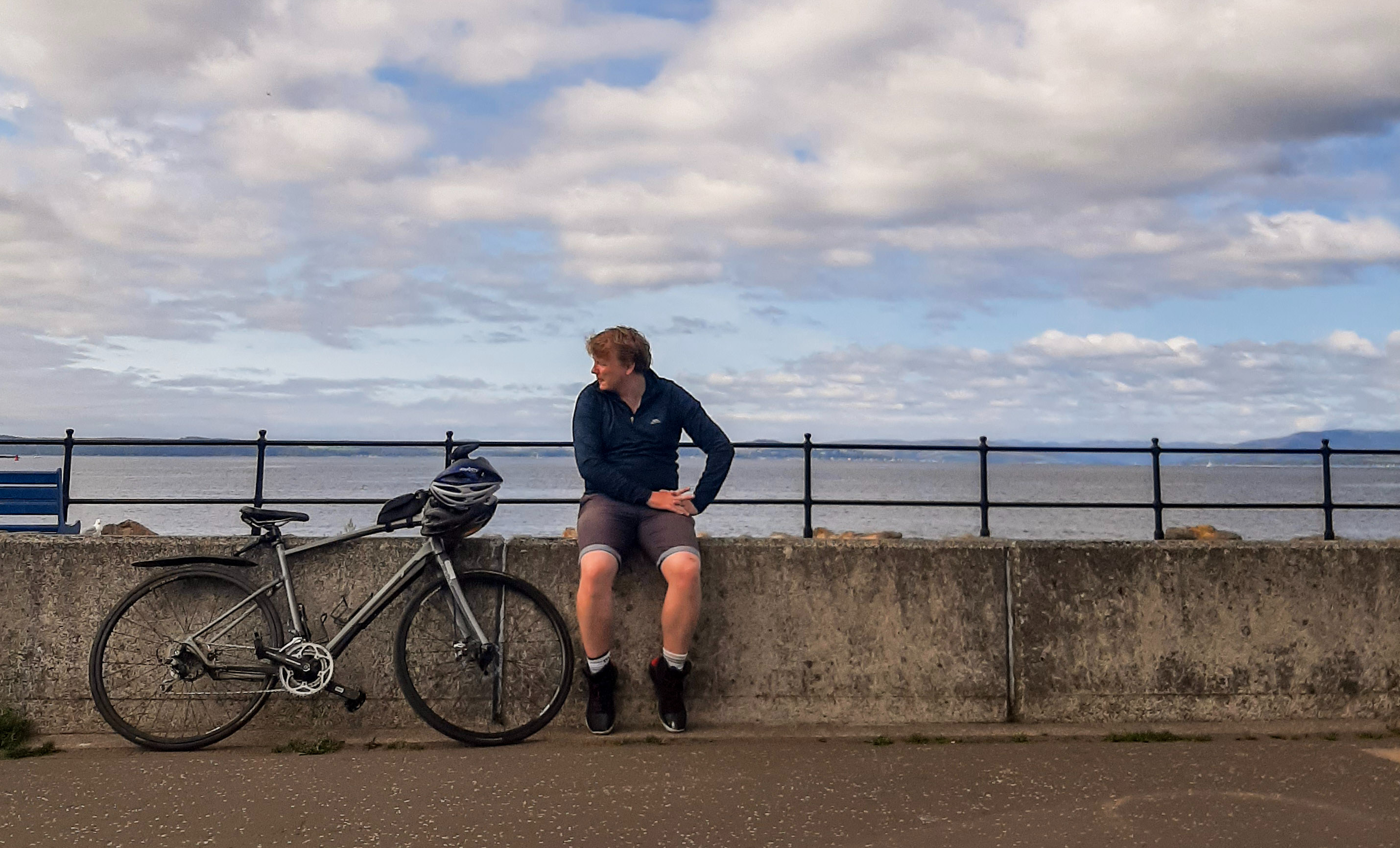 Rory is wearing a dark blbue jacket and shorts, and is sat on a concrete sea wall. To this left is a black bike, and the sea and sky are blue and white behind him.