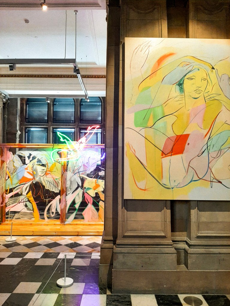 A black and white checkerboard floor on which a colourful, pastel, and neon art installation sits. In the foreground is a pastel and colourful mixed-media piece of a nude woman.