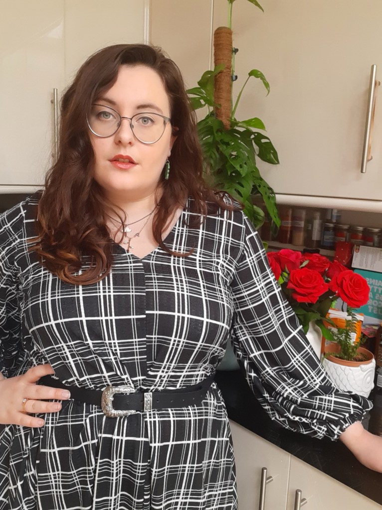 Rosie is looking at the camera and has her hand on her hip. She is wearing a long sleeve, black and white check midi dress which has a v-neck. She is also wearing a black Western belt.