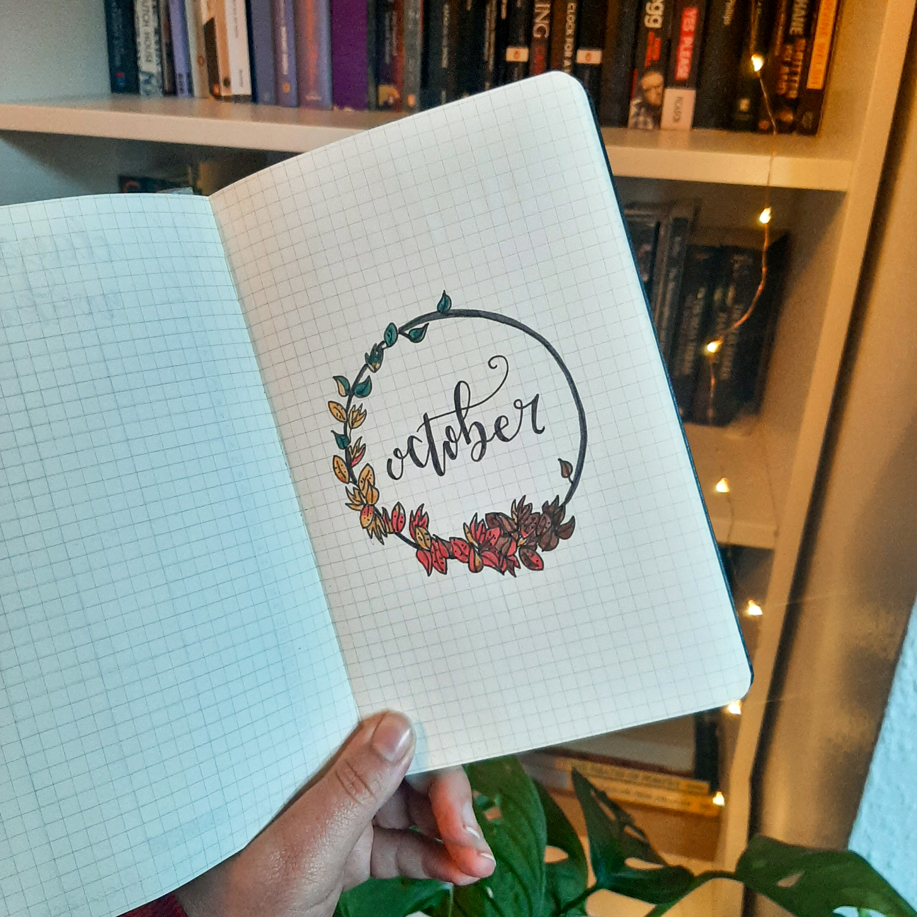 A picture of Rosie's bujo for October. The page has a black circle surrounded by leaves, with cursive 'October' written in the middle. It is held up in front of a bookcase, fairylights, and a monstera