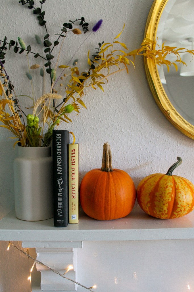 An autumnal scene on a white mantlepiece. Left-right: dried flowers of yellow and green in a white vase; a black book with 'Richard Osman, Thursday Murder Club' on the spine; a yellow book with 'Welsh Folk Tales, Peter Stevenson' in red on the spine; two small-ish pumkings, one orange, one yellow and orange. Above is a gold mirror, below are some yellow fairy lights.