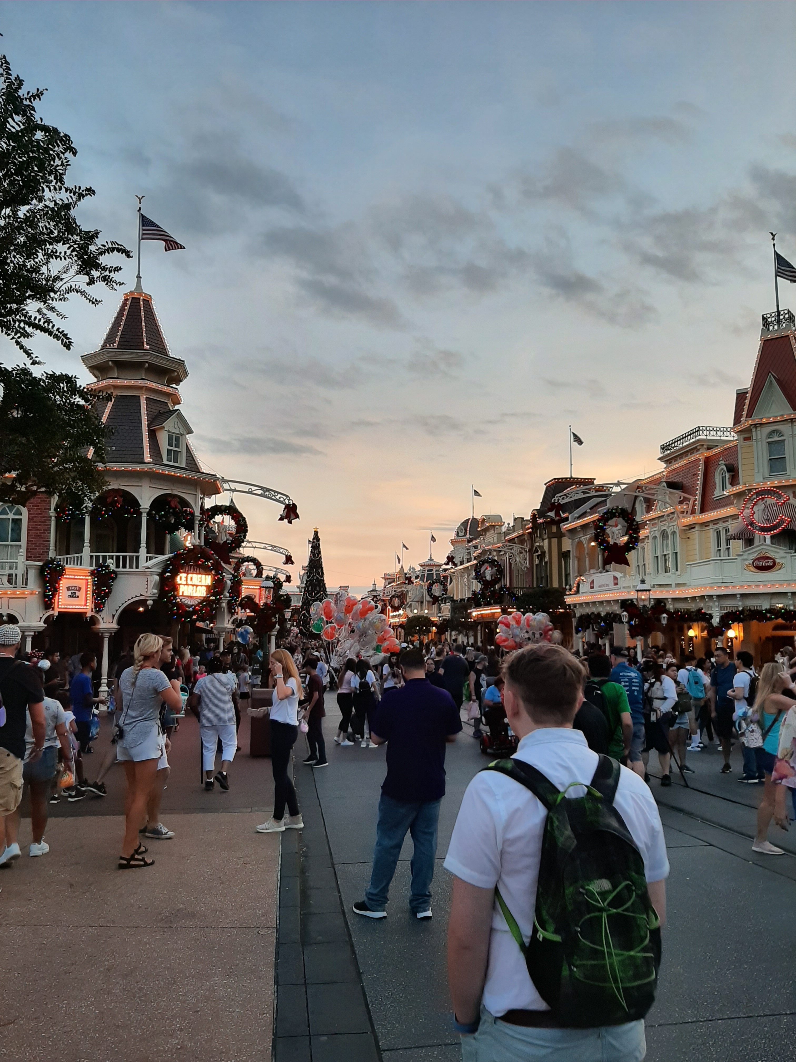 A blue sky fading into orange. Below the sky lies a dusk lit scene of Main Street USA with yellow buildings, balloons, and crowds of people. Rory is walking into the crowd, with a white shirt, and a black rucksack.