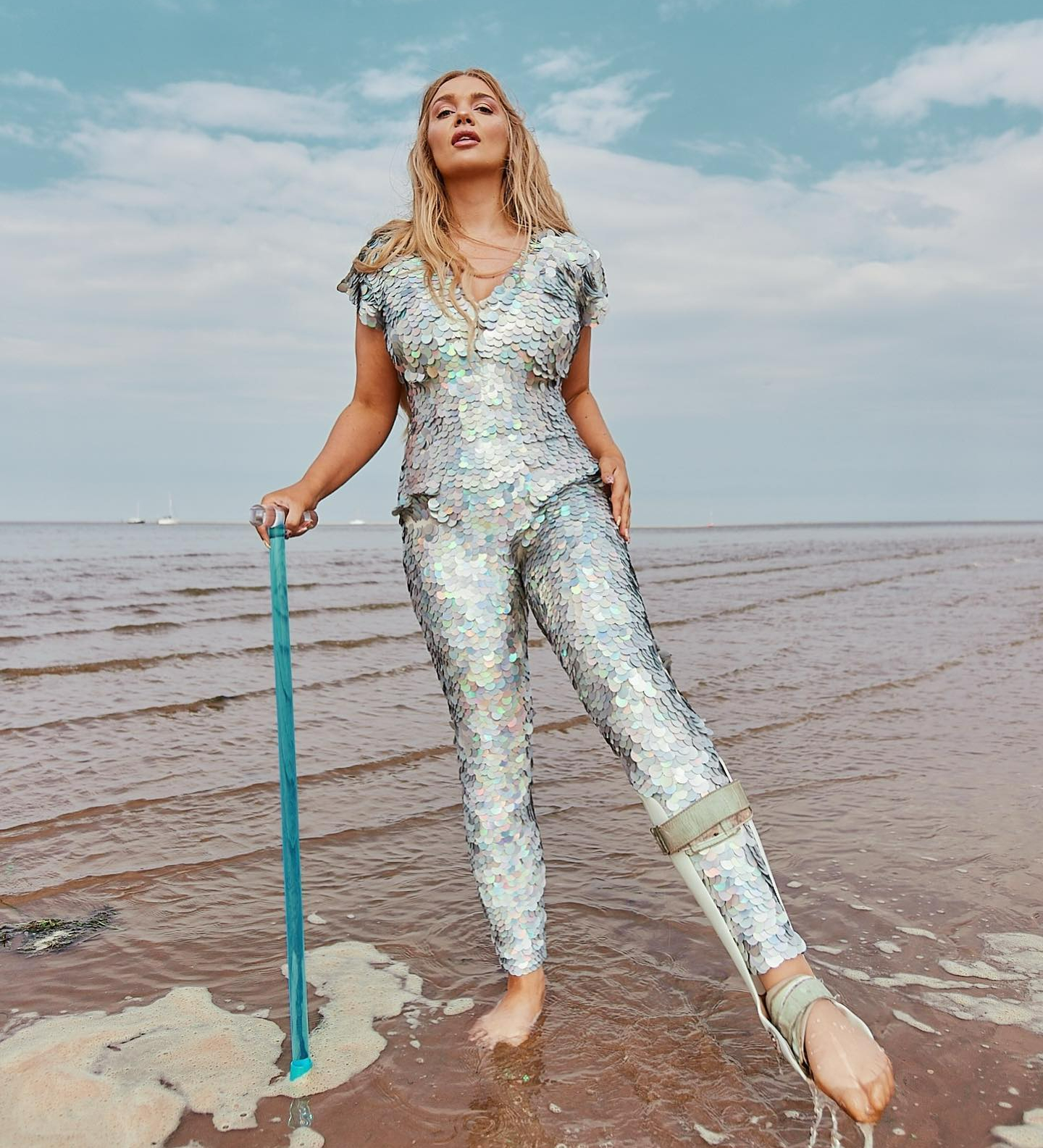 A picture of Lucy May Dawson. She is a white woman with long blonde hair. She is wearing a full body silver sequin jumpsuit and is holding a blue cane in her right hand. She is posing with her left leg pointed, and on her left leg is a splint. She is stood in the sea on the shallows, with the blue sky behind her.
