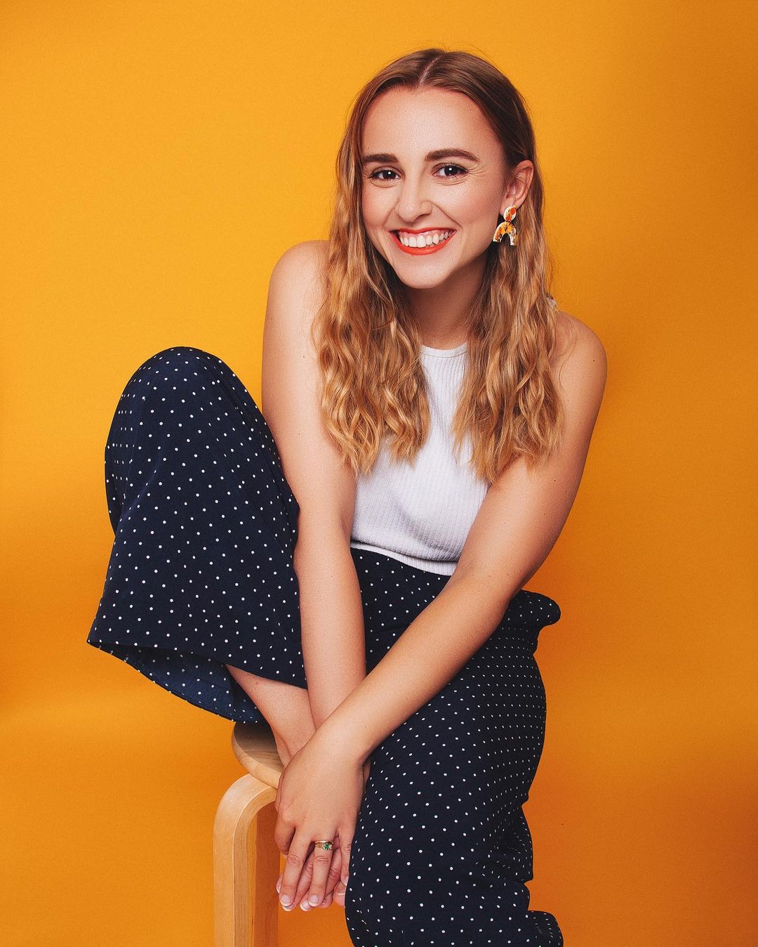 A picture of Hannah Witton. Hannah is a white woman with brown-blonde hair. She is sat on a stool with her right leg up. She is smiling at the camera, has orange lips, and curved orange earrings. She is wearing a white vest and black culottes with white dots.