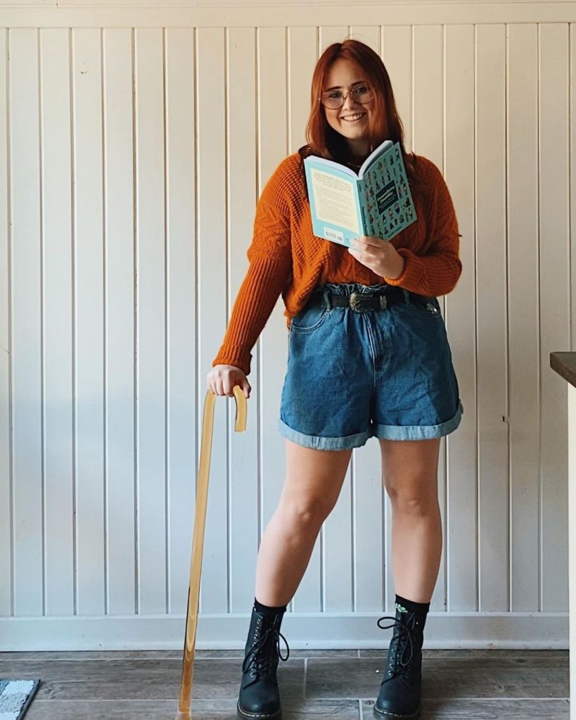Picture of Autumn from theliteraryheroine. Autumn is a white eoman with straight red hair. She is standing and using a brown cane for support on her right. In her left hand, she is holding a pale blue book. She is wearing an orange jumper, blue denim shorts, and black boots.