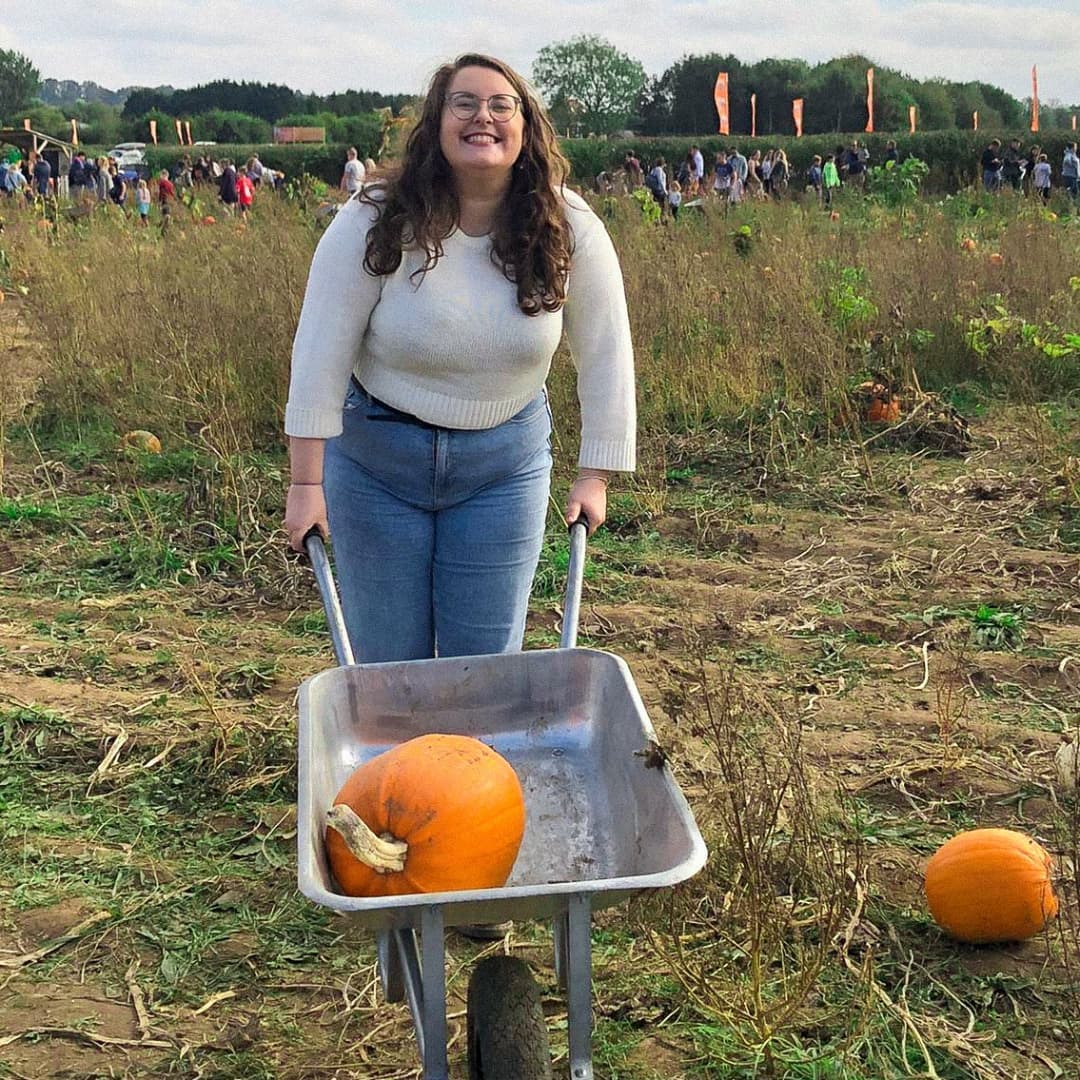 A full body picture of Rosie as she looks at the camera. Rosie is white woman with brown wavy hair. She is grinning at the camera, and is pushing a silver wheelbarrow. She is wearing a cream jumper and light blue mom jeans.