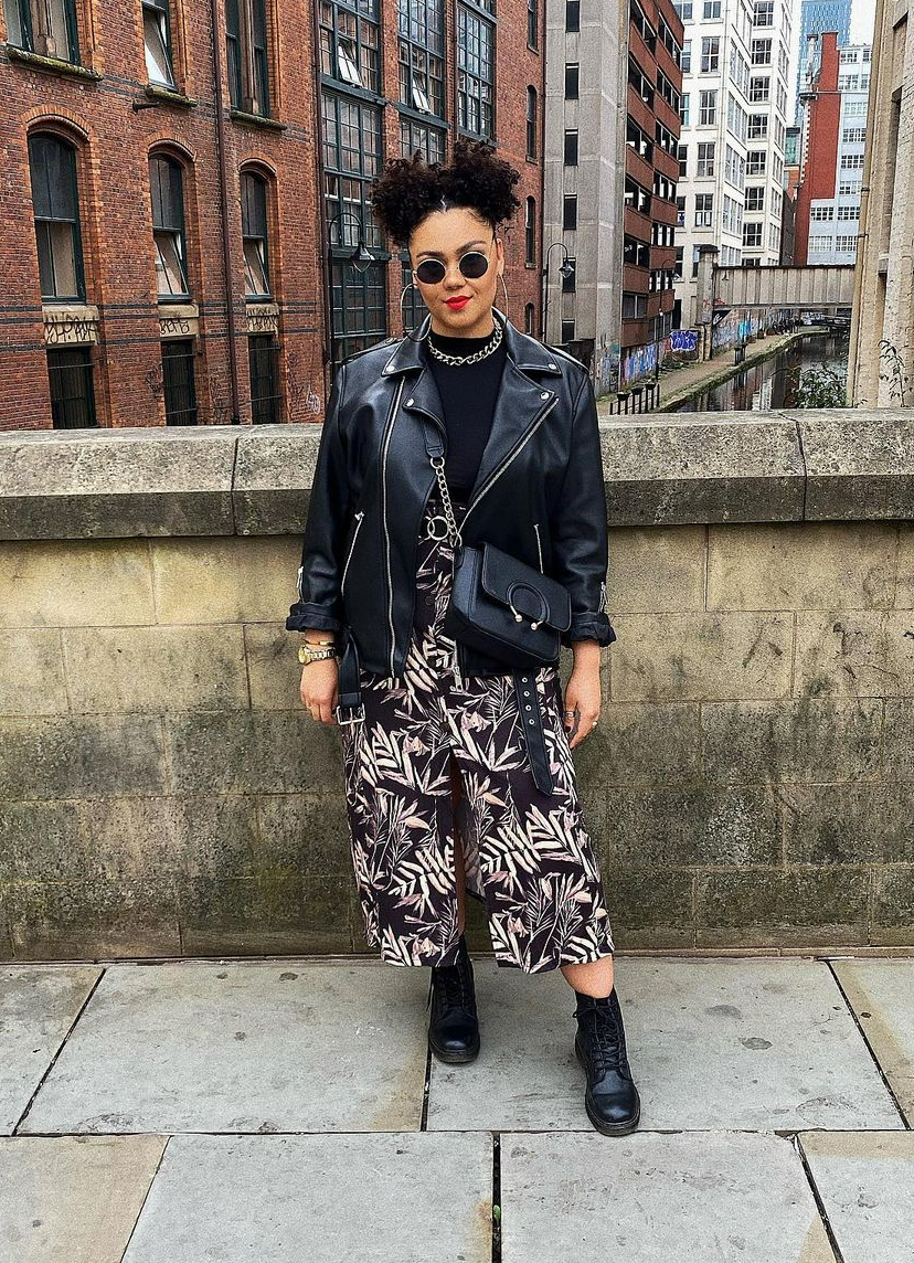 An image of Charleigh Victory. Charleigh is a light-skinned black woman, and has black curly hair in two buns. She is stood on a bridge, and is wearing black sunglasses with a red lip. She is wearing a black tee, black leather jacket, black culottes with white botanical print, and black boots.