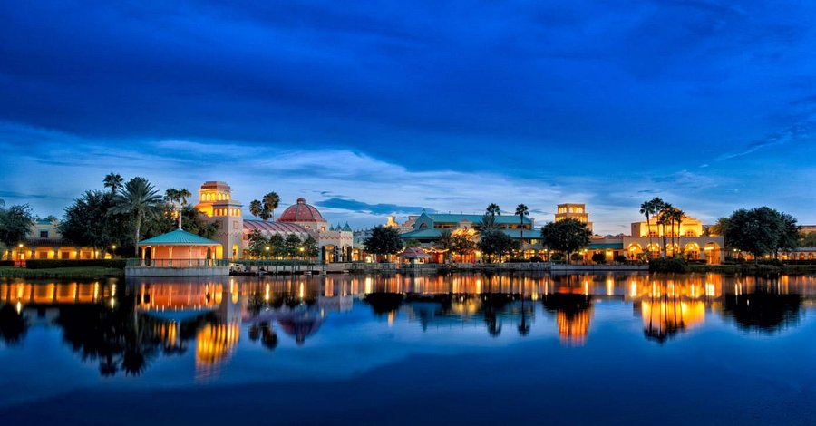 A landscape shot Coronado Springs at dusk. The sky above and the water below and both deep blue. Across the centre of the image, Mexican themed buildings of the resort shine yellow and orange, and reflect on the water. 