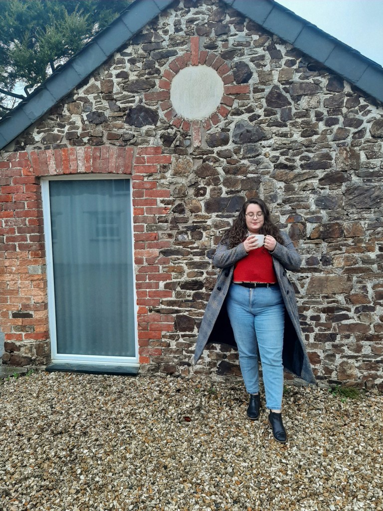 A picture of Rosie sipping out of a white coffee cup. She is wearing a red jumper, blue jeans, black boots, and a grey coat. She is Caucasian, with dark brown hair and glasses. She is stood infront of a rustic brick wall.